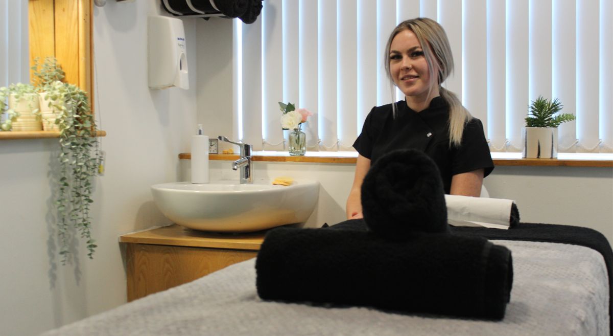 female student sat behind a treatment bed with rolled up towels in the foreground, and a sink the background